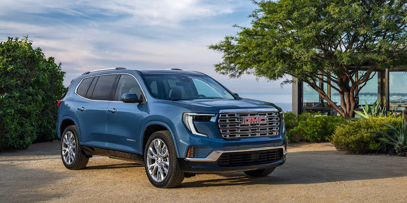 Blue GMC Acadia SUV parked outdoors near trees and a waterfront view.