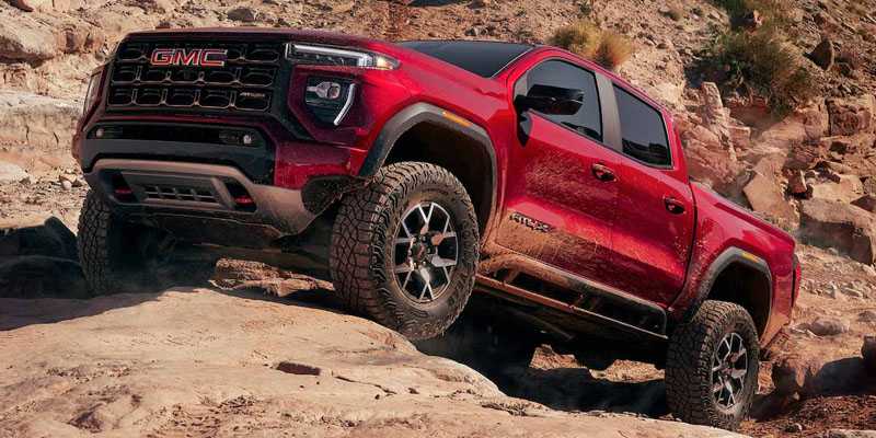 Red GMC pickup truck climbing over rocky off-road terrain in a desert landscape.